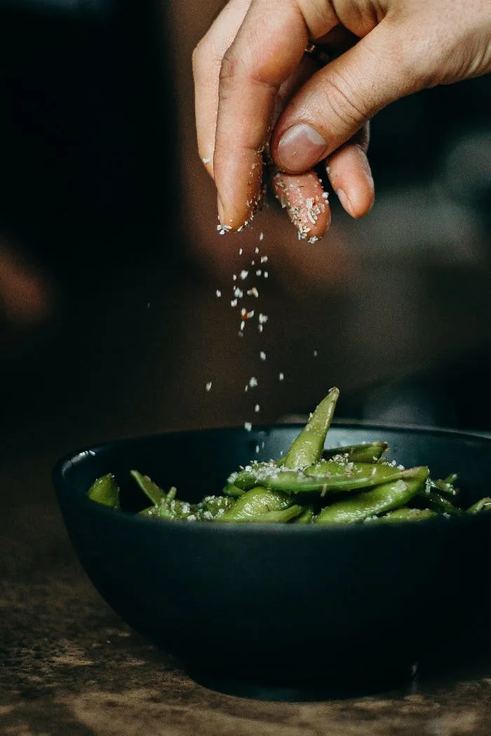 Food Image 1 A hand sprinkles salt on fresh edamame, showcasing a delicious and healthy cooking moment.